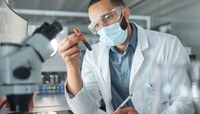 Técnico com jaleco, óculos protetor e máscara avalia amostra em laboratório.