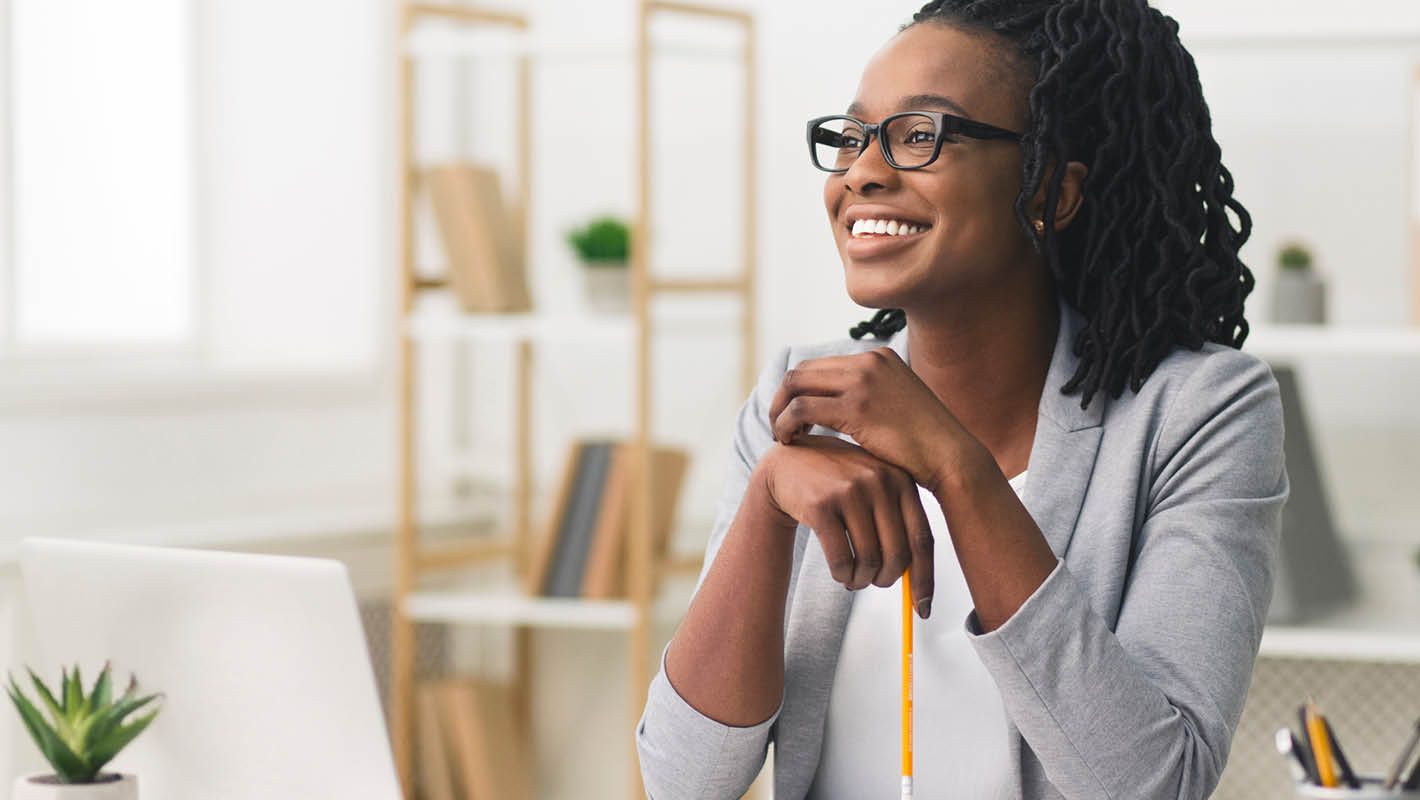 Mulher negra com tranças e roupas sociais segura lápis diante de notebook e sorri dentro de escritório