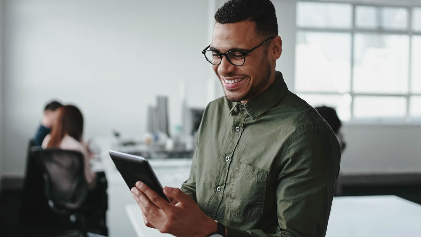 Homem negro com roupa social sorri enquanto trabalha em tablet em escritório com equipe ao fundo