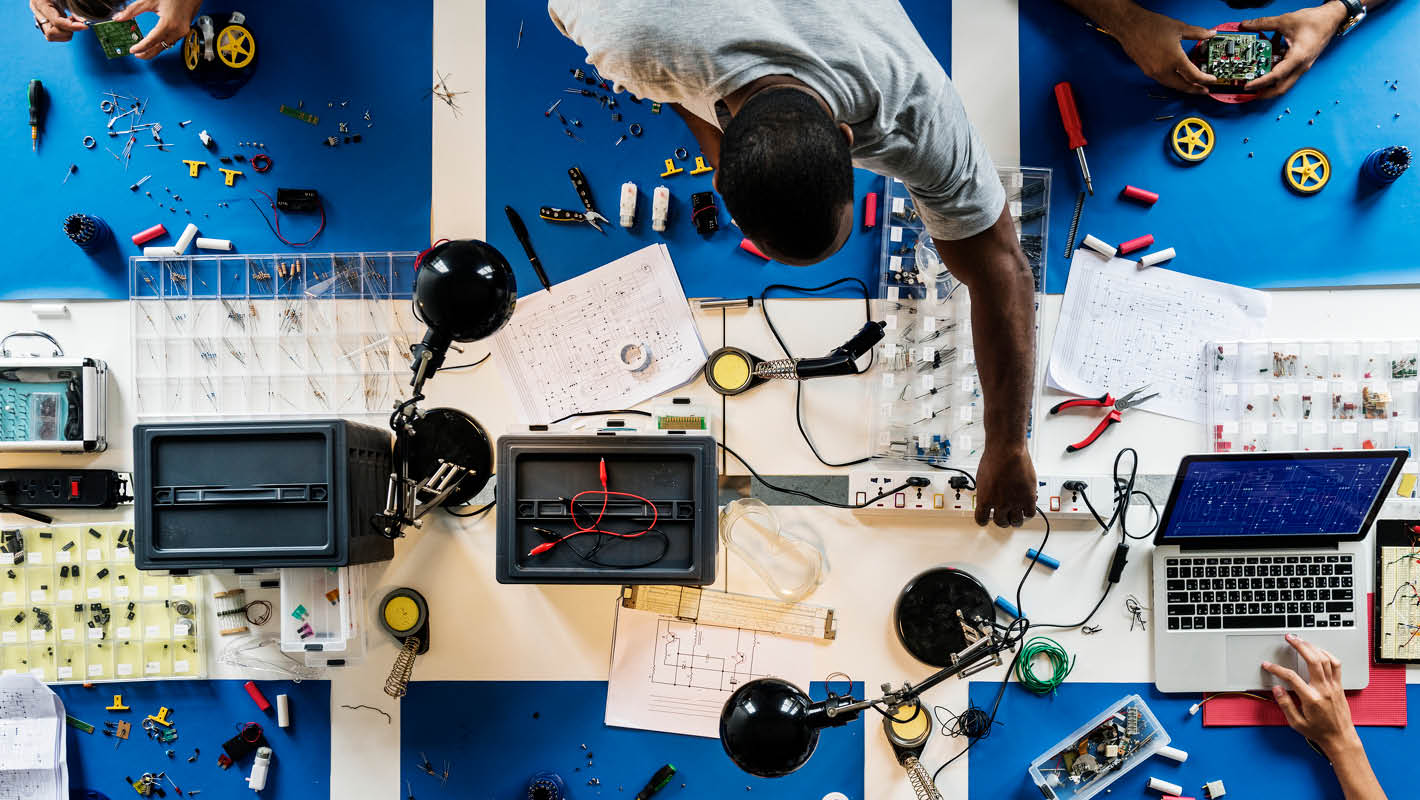 Visão de cima, seis pessoas trabalham sobre mesa, alguns olhando projetos no papel, outros no notebook, outros constroem robôs e montam circuitos.