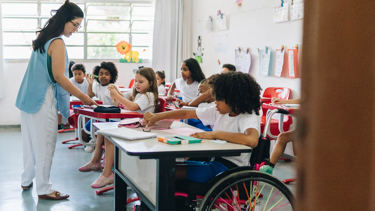 Professora diante de turma em sala de aula com a presença de estudante cadeirante.