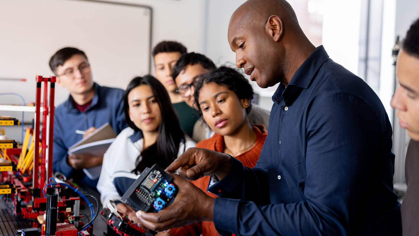 Professor dá aula para turma de estudantes e aponta para peça e componentes eletrônicos.